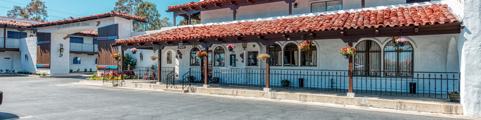 A two-story building with red-tiled roofs and arches features a parking lot in front under a clear blue sky.