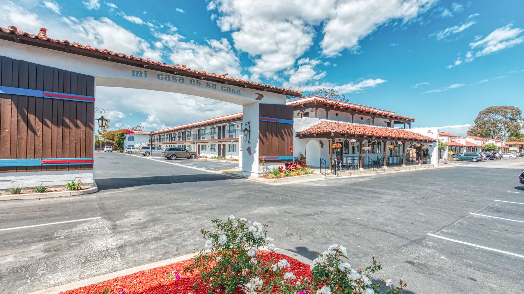 The image shows a motel with a parking lot, landscaped with red mulch and flowers, blue skies, and a white archway.