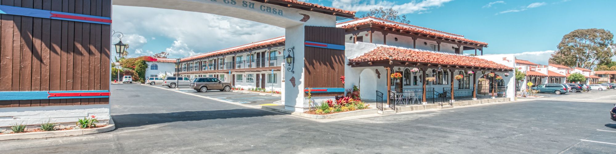 The image shows a motel with a parking lot, landscaped with red mulch and flowers, blue skies, and a white archway.