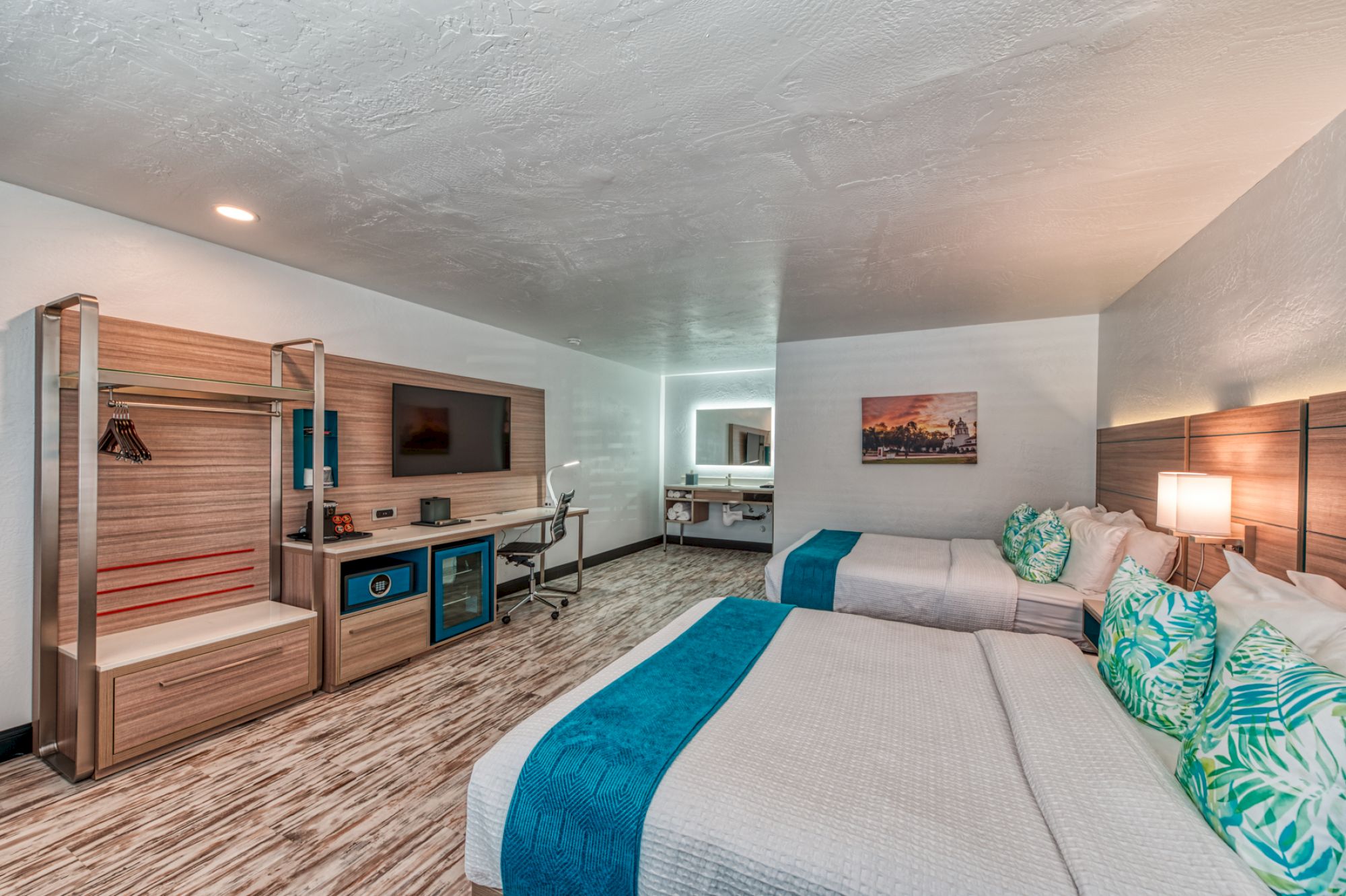 A modern hotel room with two large beds, a TV, desk, and a small kitchenette area. There are pillows with green leaf patterns on the beds.