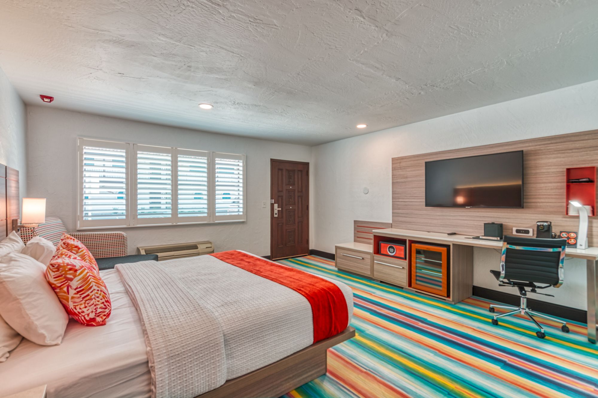 A modern hotel room with a double bed, colorful striped carpet, large TV, workspace, and window with blinds. The room is neatly organized and bright.