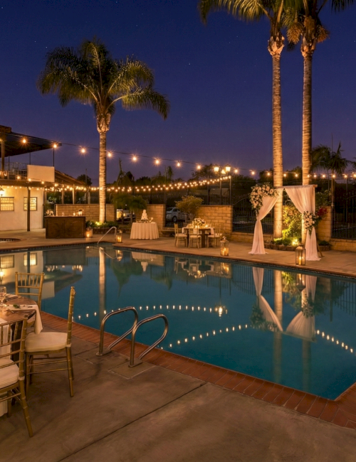 Elegant poolside evening setup with string lights, palm trees, and round banquet tables adorned with flowers around a glowing rectangular pool.