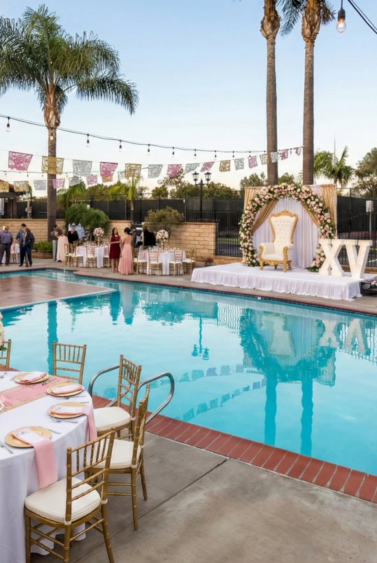 A stylish poolside party setup with round pink-tabled tables, gold chairs, floral centerpieces, string lights, and guests mingling around a bright blue swimming pool.
