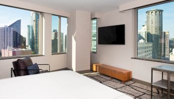 A modern hotel room with large windows offering a city view, a wall-mounted TV, a chair, a wooden bench, and a white bed.