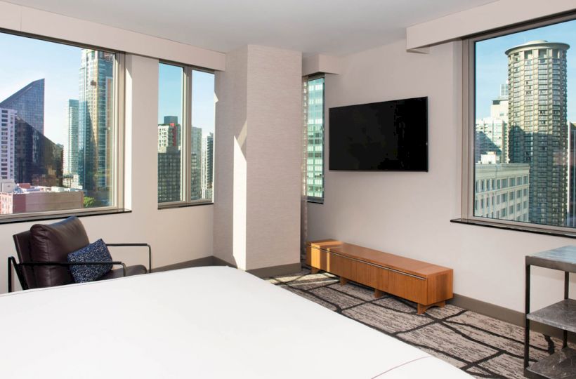 A modern hotel room with large windows offering a city view, a wall-mounted TV, a chair, a wooden bench, and a white bed.