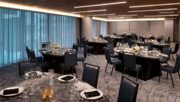 The image shows an elegantly decorated banquet hall with round tables set for a formal event, featuring dark chairs, folded napkins, and glassware.