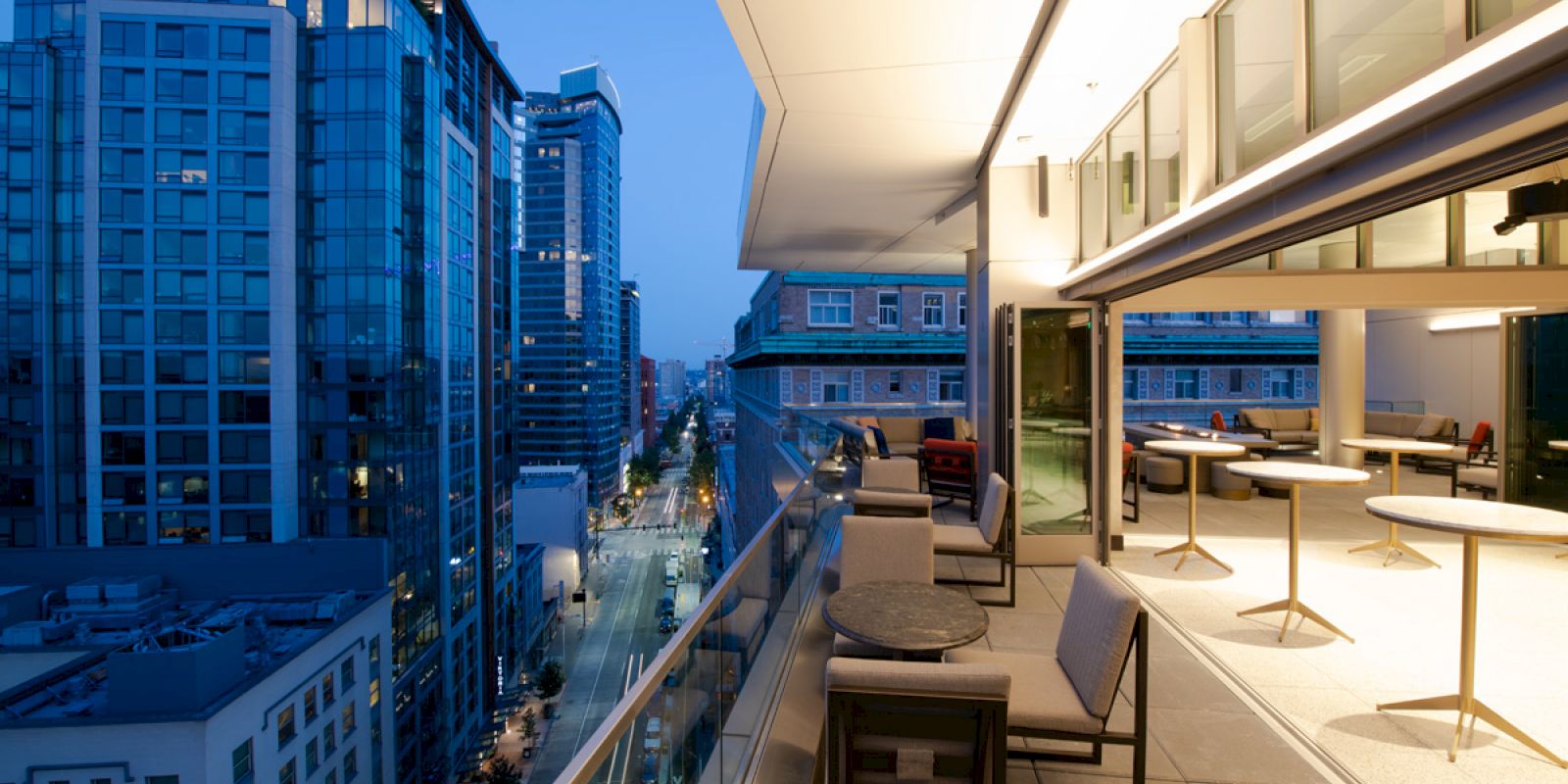 This image shows a modern cityscape with tall buildings at dusk, viewed from a balcony with seating and tables.