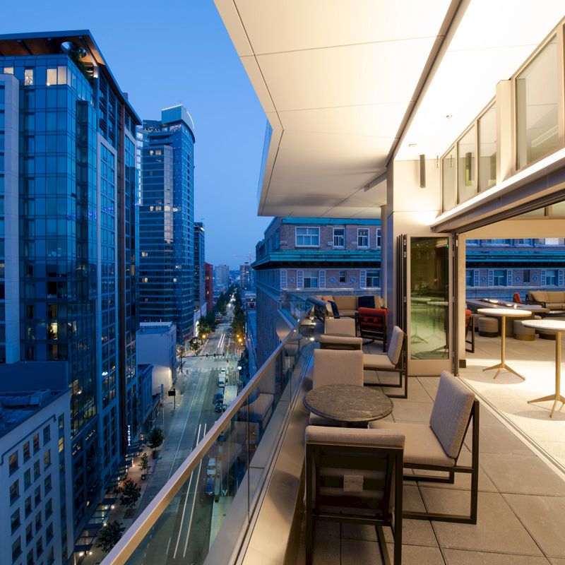 A modern urban balcony with seating and tables overlooks a city street lined with tall buildings during twilight, showcasing a vibrant cityscape.