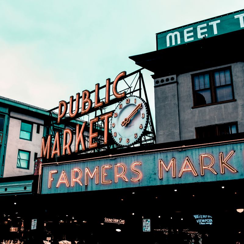 This image shows an iconic "Public Market" and "Farmers Market" sign with a large clock, set in an urban environment with surrounding buildings.