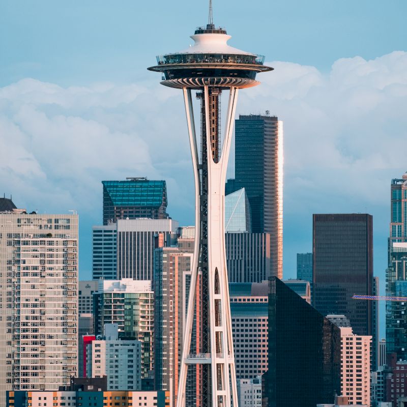 The image shows an urban skyline with the iconic Space Needle tower prominently featured among various modern buildings and skyscrapers under a clear sky.