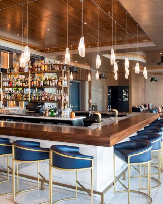 The image shows a stylish, modern bar with a wooden countertop, blue bar stools, hanging lights, and a wide variety of bottles on display.