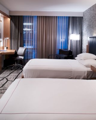 A modern hotel room with two neatly made beds, a wall-mounted TV, a work desk with a chair, and stylish lighting and decor.