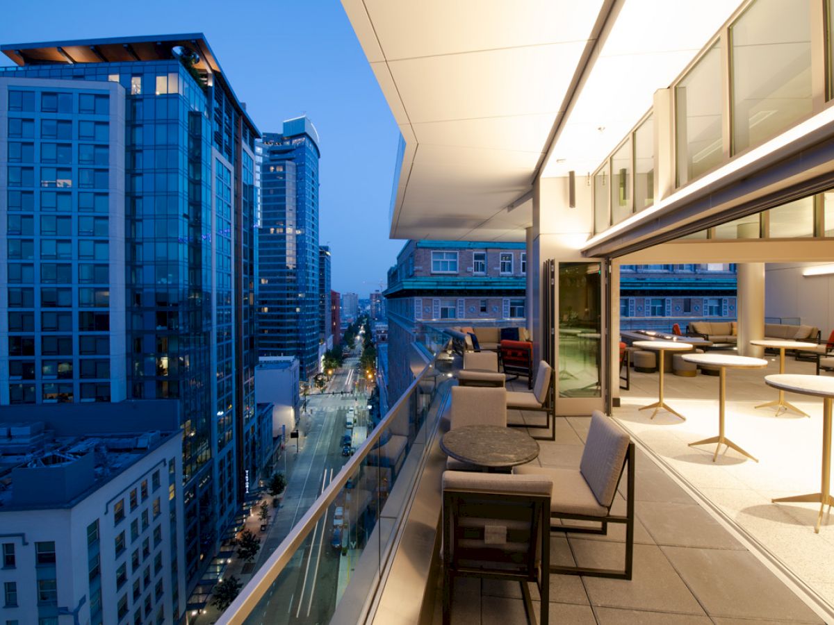 A modern balcony at dusk with seating and tables, view of a city street, and high-rise buildings in the background.