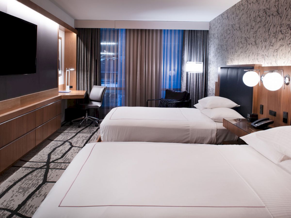 This image shows a modern hotel room with two neatly made beds, a desk with a chair, a wall-mounted TV, and a well-lit window.
