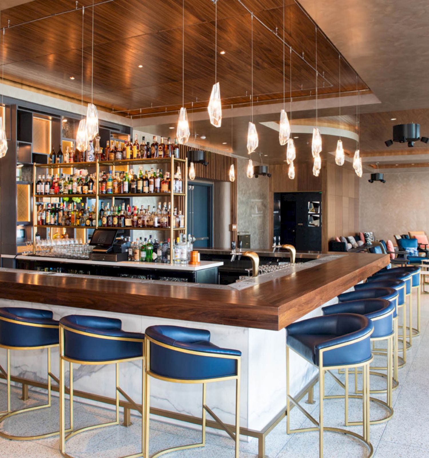 The image shows a modern bar with blue chairs, a well-stocked liquor shelf, hanging light fixtures, and a spacious seating area with couches.