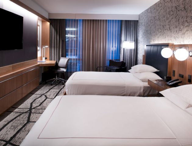 This image shows a modern hotel room with two beds, a large TV, a desk with a chair, and stylish wall decor and lighting.