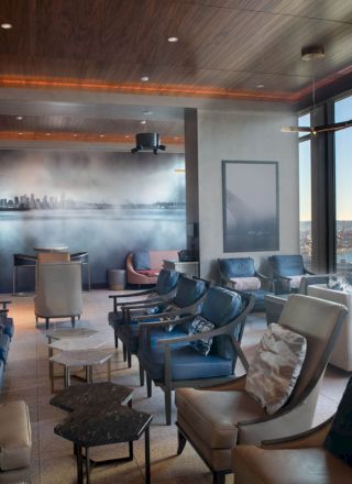 The image shows a modern lounge area with numerous chairs, tables, and large windows offering a scenic view of a bay and a Ferris wheel outside.
