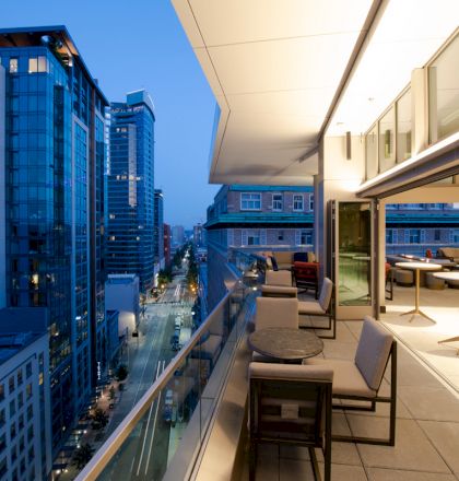 The image shows a modern balcony or terrace overlooking a city street with tall buildings, furnished with chairs and tables, during dusk.