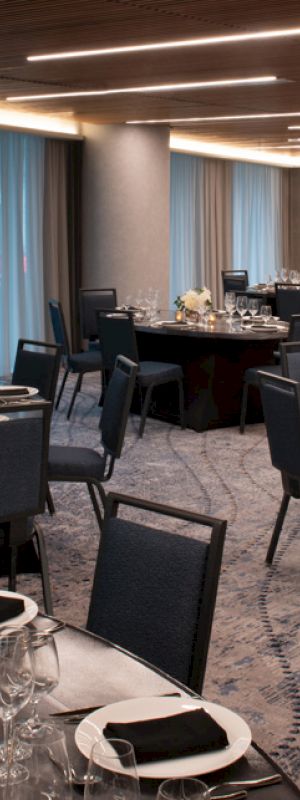 A modern event space with round tables set for dinner, featuring elegant tableware, black napkins, flower centerpieces, and dim lighting.