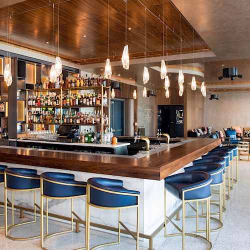 A modern bar area with a wooden counter, blue barstools, a variety of spirits on shelves, pendant lights, and a lounge seating area in the background.