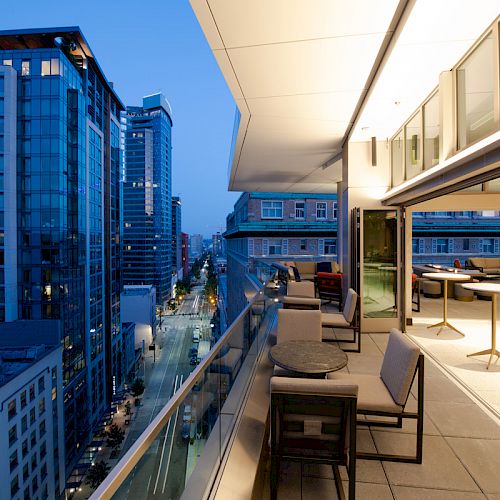 This image depicts a modern cityscape at dusk, viewed from a balcony with outdoor seating, overlooking a street with high-rise buildings.
