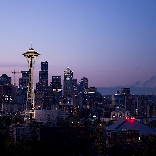 A city skyline at dusk with the Space Needle prominently visible, Mount Rainier in the background, and skyscrapers illuminated against the evening sky.