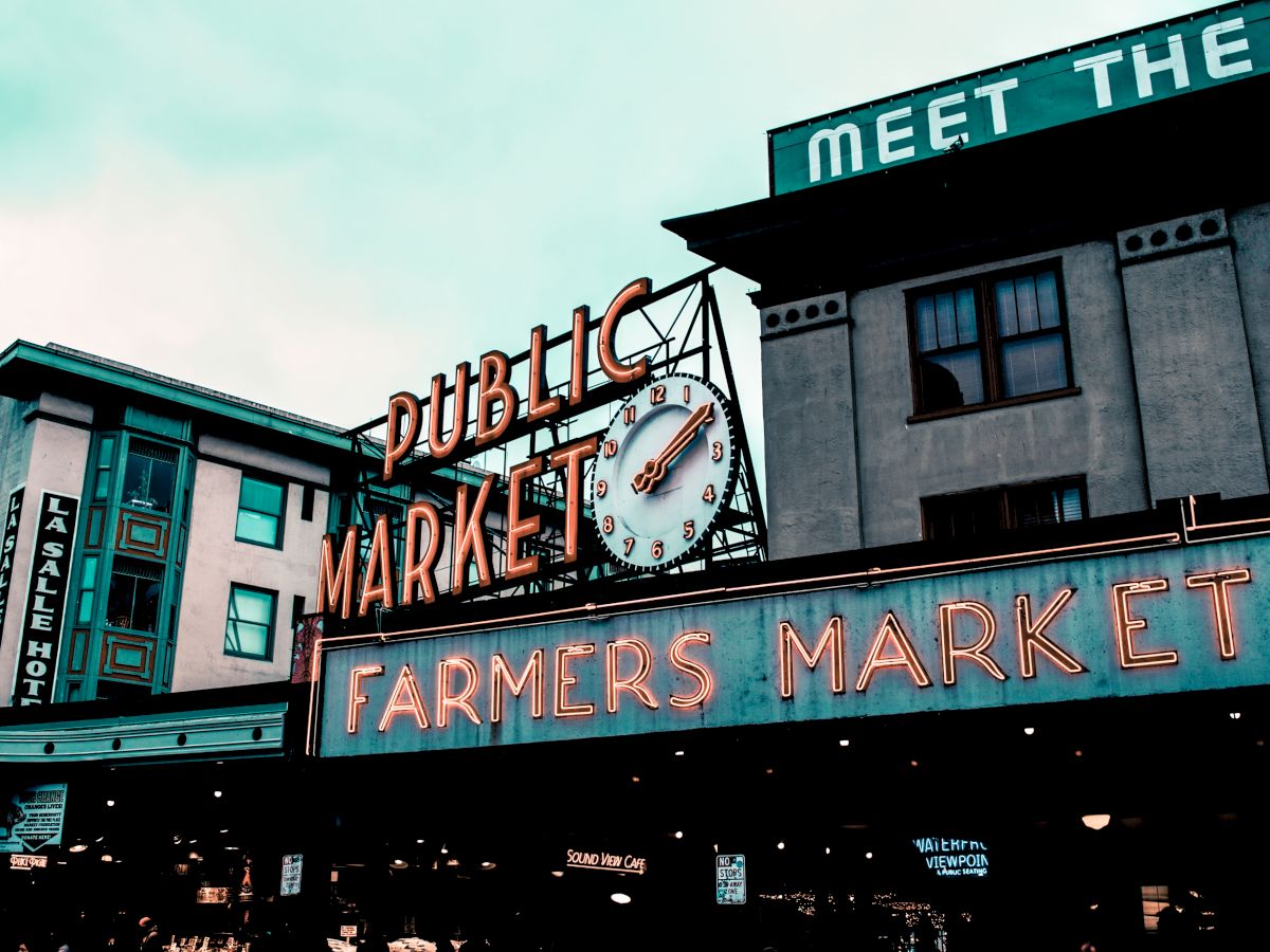 The image shows the famous "Public Market" and "Farmers Market" signs at Pike Place Market in Seattle, Washington, known for its vibrant marketplace.