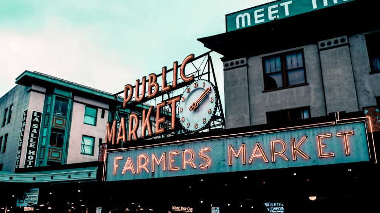 The image shows the famous "Public Market" and "Farmers Market" signs at Pike Place Market in Seattle, Washington, known for its vibrant marketplace.