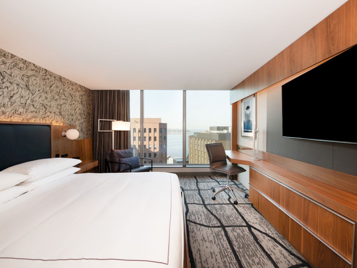 A modern hotel room with a large bed, stylish decor, a desk with a chair, a flat-screen TV, and a window offering a cityscape view.
