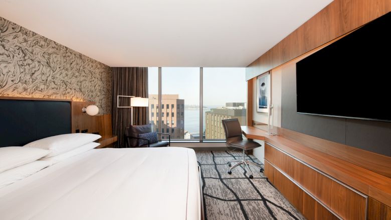 A modern hotel room with a large bed, stylish decor, a desk with a chair, a flat-screen TV, and a window offering a cityscape view.