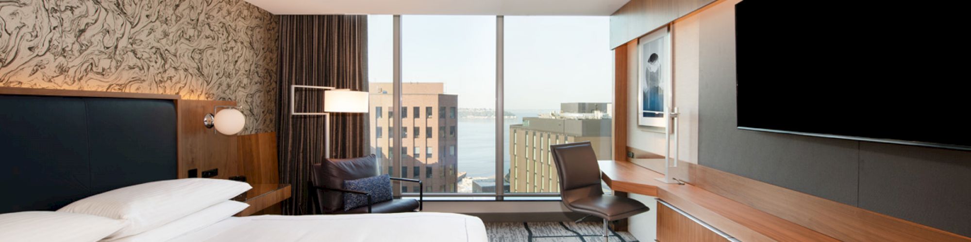 A modern hotel room with a large bed, a desk and chair, a large TV, and a window with a city view.