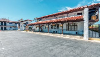 The image shows a two-story building with Spanish-style architecture, featuring arched windows, red-tiled roof, and an open parking lot in front.